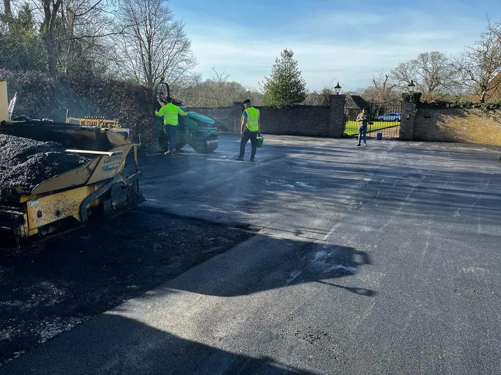 A paver and roller laying a large area of fresh tarmac, the crew working across a wide surfaced forecourt.