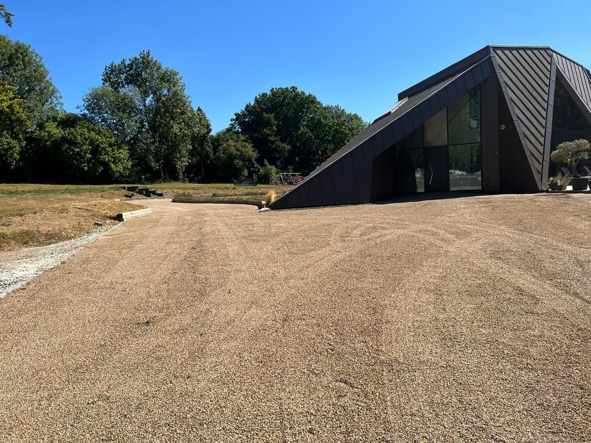 A broad, evenly raked golden-gravel driveway sweeping up to a modern architect-designed house with dark angular cladding and a large glazed gable.