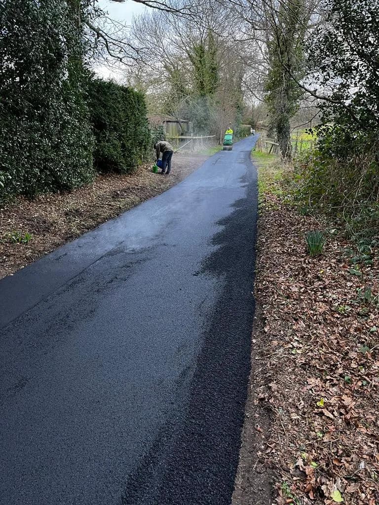 A freshly surfaced rural lane in smooth black tarmac, running between hedgerows.